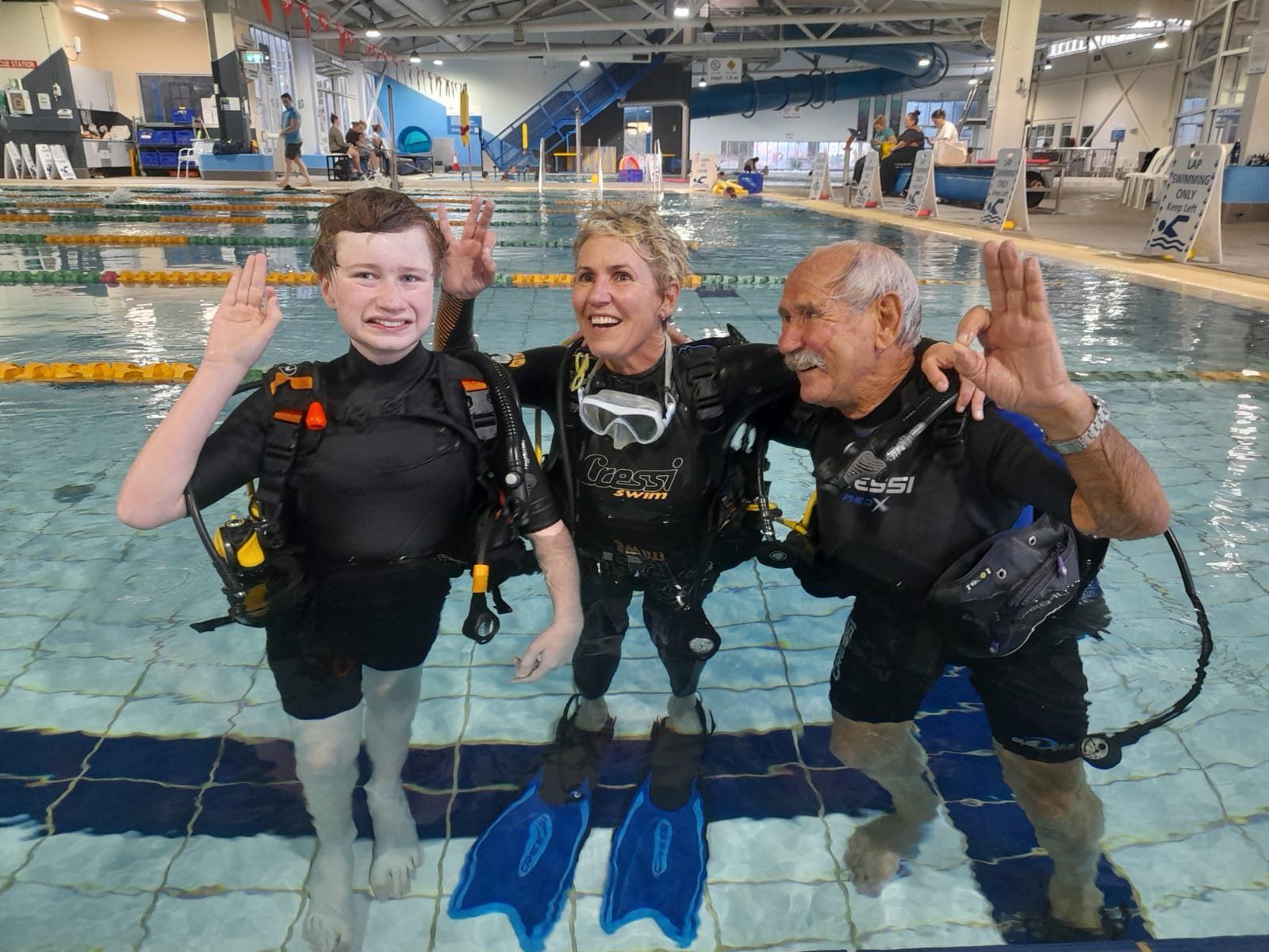 Three divers in a pool with fins, giving the OK sign